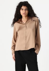 Lichtbruin satijnen overhemd met een puntige kraag, knoopsluiting en lange mouwen. Glad textuur met een ontspannen pasvorm. Zwarte broek eronder.