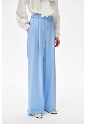 Kobieta ubrana w jasnoniebieskie spodnie z wysokim stanem i szerokimi nogawkami z zakładkami, zestawione z białą bluzką i białymi szpiczastymi butami z ćwiekami.