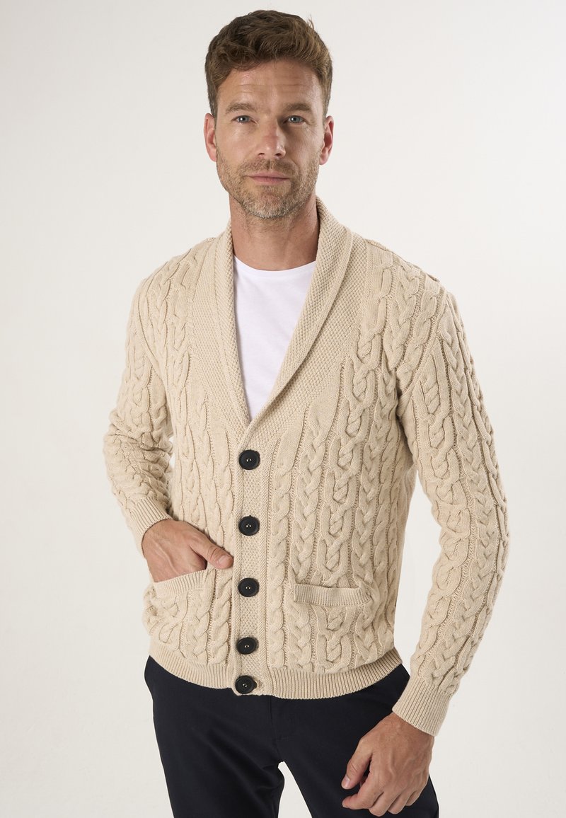 Beige kabelstrik cardigan med sjalskrave, der har sorte knapper og frontlommer; den teksturerede design tilføjer visuel interesse.