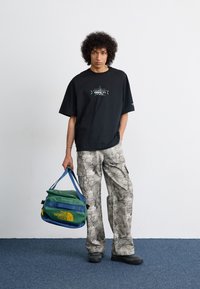 Black oversized T-shirt with front graphic, light gray camouflage cargo pants, and a green/blue The North Face duffle bag. Black shoes.