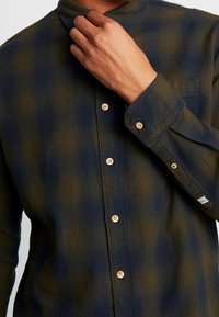Camicia button-up in plaid blu navy e verde oliva, realizzata in tessuto morbido. Presenta un colletto a punta e bottoni in legno con una tasca sul petto.