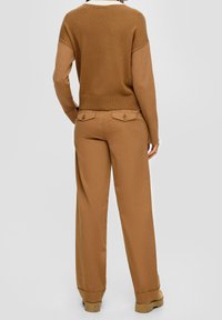 Personne portant un pull coloré marron et un pantalon large beige avec des poches arrière à boutons, se tenant avec les mains dans les poches.