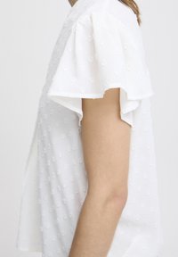 Blusa bianca e strutturata con maniche corte e volant, caratterizzata da un motivo a pois e una vestibilità rilassata. La vista laterale mette in evidenza i dettagli delle maniche.