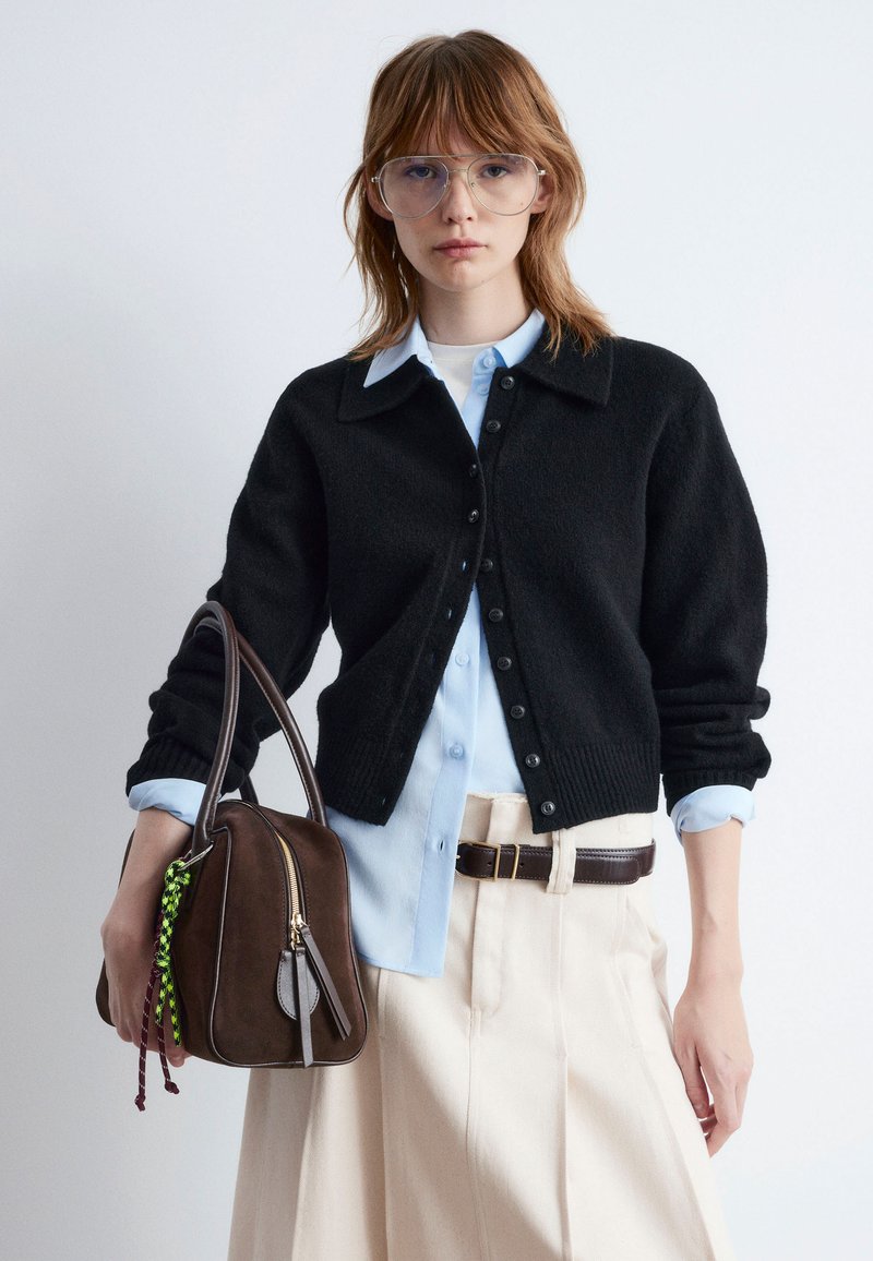 Schwarze Strickjacke, hellblaues Kragenshirt darunter. Das Model hält eine braune Lederhandtasche mit Reißverschlüssen und grünen sowie kastanienbraunen Akzenten.