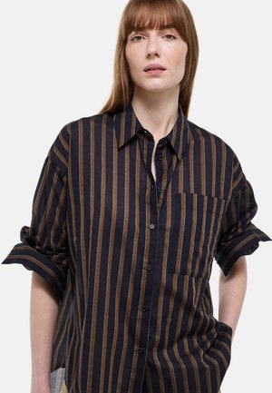 Femme portant une chemise ample à rayures verticales noires et marron, boutonnée, avec les manches retroussées, sur un fond clair uni.