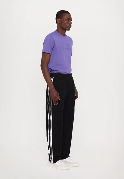 Lilla t-skjorte med korte ermer og preget logo, kombinert med svarte joggebukser med hvite sidestreker og hvite sneakers.
