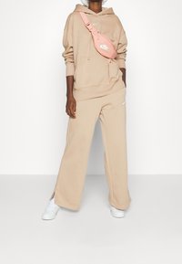 Beige hoodie och vida sweatshirtbyxor med Nike-logotyp. En rosa axelremsväska vilar på axeln. Vita sneakers fulländar outfiten.