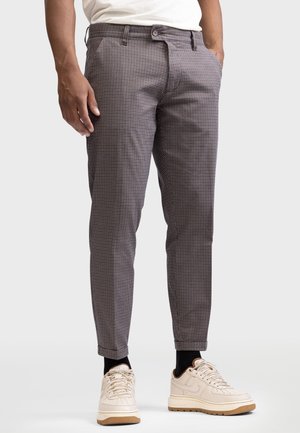Man die een slim-fit geruite broek, beige sneakers met zwarte sokken en een wit overhemd draagt, staand met één hand in zijn zak.