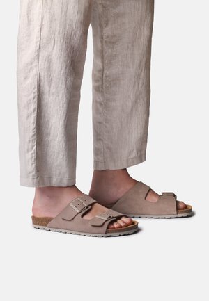 Nohy v béžových semišových pantoflích na nazutí se dvěma nastavitelnými přezkami, doplněné o světle béžové zkrácené lněné kalhoty.