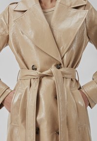 Beige, skinnende faux læder trench coat med en dobbeltradet design, brede klapper og en bindebælte i taljen. Har to sidelommer.