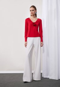 Rotes langärmliges Oberteil mit V-Ausschnitt und Blumendetail in der Mitte, kombiniert mit weißen weitem Hosen und schwarzen Schuhen.