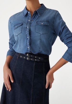 Kobieta ubrana w niebieską, dżinsową koszulę na guziki z kieszeniami na piersi, wpuszczoną w ciemną dżinsową spódnicę z podwójnym paskiem z klamrami, ręce opierające się na biodrach.