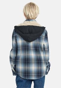Camicia in flanella a quadri nei toni del blu con cappuccio scuro e rivestimento in pelliccia sintetica. Vestibilità rilassata, maniche lunghe e polsini con un solo bottone.