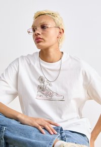 T-shirt en coton blanc avec un graphisme d'une sneaker New Balance ornée de fleurs, accompagnée d'un pantalon en denim bleu et d'un collier en chaîne argenté.