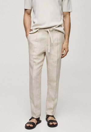 Man die beige linnen broek met trekkoorden en zwarte sandalen draagt, staand met handen in de zakken tegen een effen achtergrond.