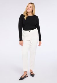 Žena v černém dlouhém rukávu se zlatými knoflíky na manžetách, bílých elegantních kalhotách a černých špičatých slingback balerínách, stojící na bílém pozadí.