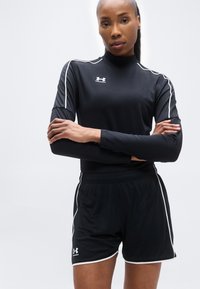 Γυναίκα με σταυρωμένα τα χέρια, ντυμένη με μαύρα αθλητικά ρούχα Under Armour με λευκές λεπτομέρειες, σε απλό ανοιχτό φόντο.