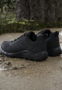 Czarne buty sportowe mają teksturowaną syntetyczną cholewkę, wyraźny wzór w trzy paski, grubą gumową podeszwę oraz wzmocnioną pętelkę przy pięcie dla łatwego zakładania.