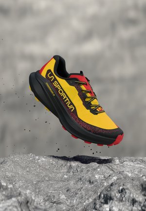 Gelber, roter und schwarzer La Sportiva Prodigy Trailrunning-Schuh schwebt über felsigem Gelände mit kleinen herabfallenden Steinen.