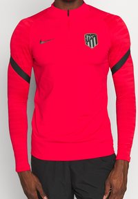 Top deportivo de Nike en rosa brillante con mangas largas y un cuello con media cremallera. Presenta líneas de acento negras y un logo del equipo en el pecho.