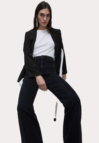 Svart kavaj över en vit t-shirt och högmidjade mörka jeans. Modellen har långt hår och bär glittrande örhängen. Sitter på en stol.