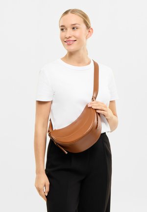 Geantă de umăr din piele maro cu un design curbat, curea unică și închidere cu fermoar. Asortată cu un tricou alb și pantaloni negri.