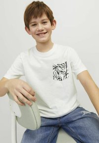 White cotton t-shirt with short sleeves, featuring a black floral-patterned pocket. Model wearing blue jeans, seated on a light-colored chair.