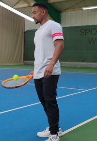 Grå t-skjorte med korte ermer og rosa striper, sorte bukser, holder en oransje tennisracket med en grønn ball på en blå bane.