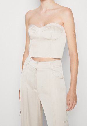 Frau trägt ein trägerloses cremefarbenes Bustier-Oberteil, kombiniert mit hoch taillierten cremefarbenen Hosen, stehend vor einem schlichten hellen Hintergrund.