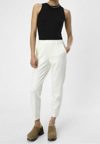 Top sin mangas de canalé negro, pantalones blancos a medida con pliegues frontales y zuecos marrones con tachuelas plateadas en un tacón de madera.