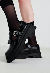Des chaussures à plateforme chunky noires avec des boucles, portées avec des chaussettes noires transparentes, les jambes croisées contre un fond clair.