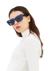 Occhiali da sole oversize blu con montatura angolare e lenti verdi, abbinati a un top bianco a collo alto. Finitura opaca con bracci neri.
