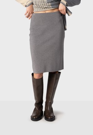 Personne portant une jupe grise longueur genou, des bottes marron montantes et un pull à motifs soulevant légèrement le pull.
