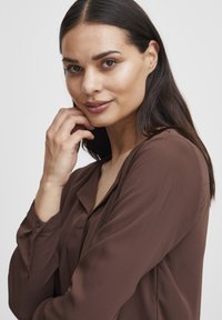 Blusa marrone a maniche lunghe in un tessuto morbido e liscio. Presenta un polsino con bottoni, un colletto classico e una vestibilità rilassata con una leggera lucentezza.