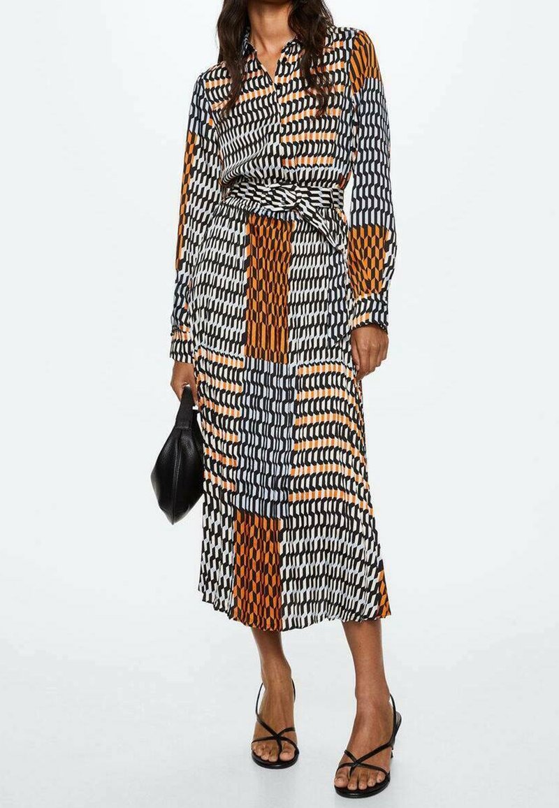 Femme en robe midi à manches longues boutonnée avec des motifs géométriques noirs, blancs, orange et gris, tenant un sac à main noir et portant des sandales noires.
