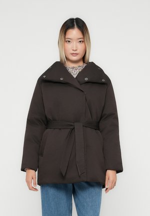 Kvinde iført en sort beltejakke i dun over en mønstret sweater og blå jeans, stående foran en ensfarvet hvid baggrund.