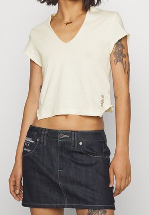 Jasnożółta, krótka koszulka z dekoltem w serek wykonana z miękkiego materiału, zestawiona z ciemnoniebieską mini spódnicą dżinsową z zielonymi przeszyciami i widocznymi kieszeniami.