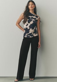 Asymmetrisches Top in Beige und Schwarz mit Batikmuster und Raffung auf einer Seite, kombiniert mit hochgeschnittenen schwarzen weiten Hosen und schwarzen Absatzsandalen.