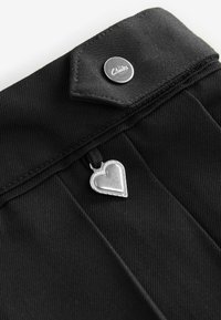 Tissu noir avec une finition texturée, doté d'un bouton argenté portant une marque et d'un charme en forme de cœur en argent attaché par une boucle noire.