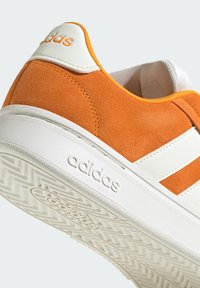 Zapatilla Adidas de ante naranja y blanco con logo en relieve en la suela y el talón blancos, que presenta detalles de marca en naranja.