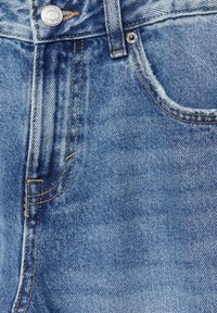 Gros plan sur un jean en denim bleu montrant la poche avant, le bouton en métal, le rivet et les détails de couture orange visibles.