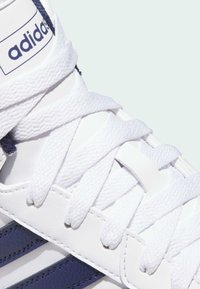 Adidas-Sneaker mit weißem strukturiertem Lederobermaterial, weißen Schnürsenkeln und blauen Streifen an der Seite. Auffälliges Branding auf der Zunge.