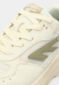 Svijetlo bež tenisica s mrežastim i brušenim materijalima, krem vezicama i zelenim logotipom s naglaskom sa strane. Teksturirani potplat za bolji prijam.