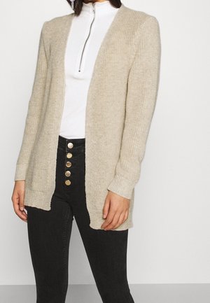 Femme portant un cardigan en maille beige ouvert sur le devant, un top blanc zippé et un pantalon noir taille haute avec cinq boutons dorés.