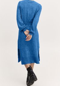 Robe tricotée bleu à manches longues, en tissu texturé, avec une taille ceinturée, des fentes latérales et des poignets côtelés. Portée avec des bottines noires à semelles épaisses.