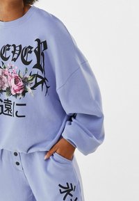 Sweatshirt en lavande en tissu doux, avec un texte noir en gras et des graphiques floraux. Manches longues et coupe décontractée avec des poignets côtelés.