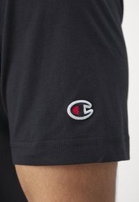 Schwarzes Kurzarmhemd aus Baumwolle. Verfügt über ein gesticktes Logo mit roten und weißen Akzenten in der Nähe des Ärmels. Glatte Textur.