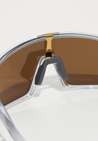 Óculos de sol com armação transparente, lentes castanhas e uma dobradiça de metal dourado. Apresentam um nariz emborrachado cinza para conforto e estabilidade.