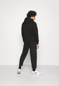 Schwarzer Kapuzenpullover mit Kapuze, kombiniert mit schwarzen Cargo-Hosen mit Seitentaschen. Weiße Sneaker mit schwarzen Akzenten runden den Look ab.