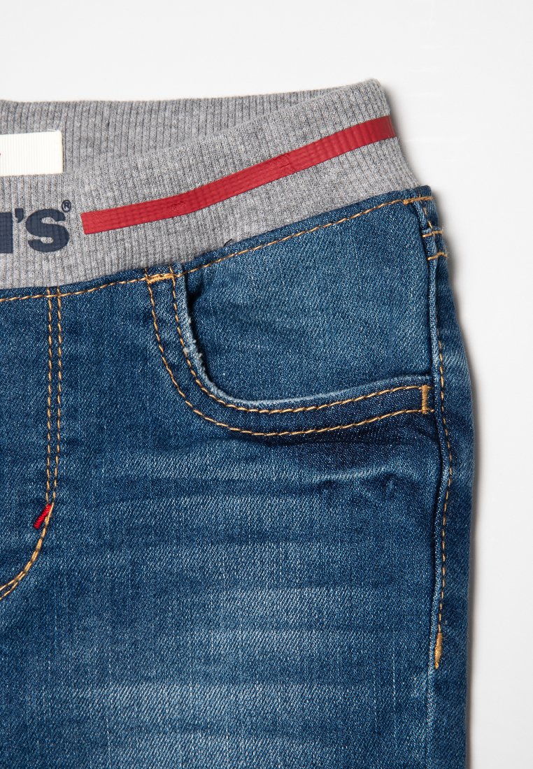 Pantalones vaqueros de mezclilla con acabado azul, que presentan una cinturilla gris con una franja roja. Incluyen costuras visibles y un pequeño bolsillo frontal.